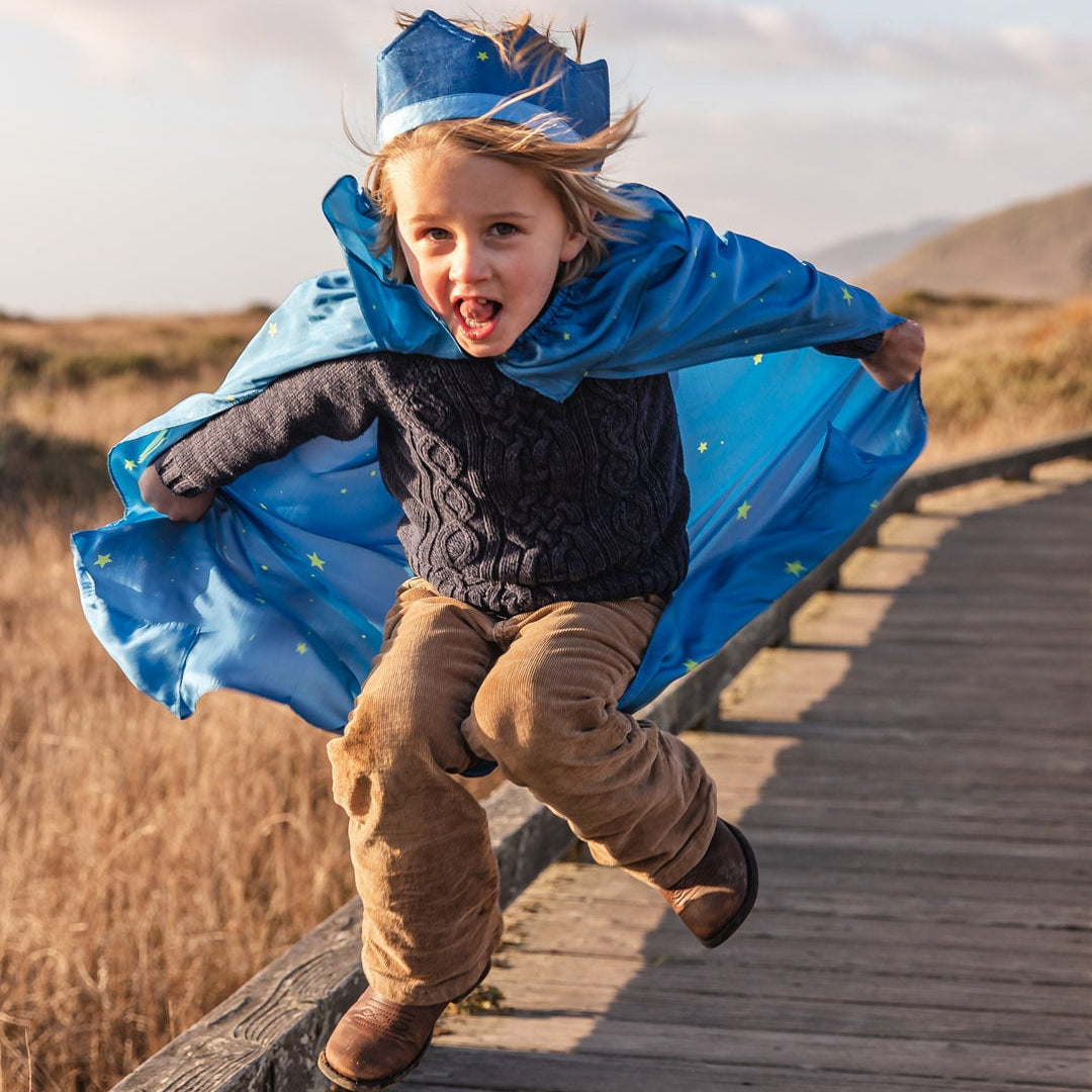 A young child wearing a blue crown and cape runs excitedly along a wooden boardwalk outdoors.