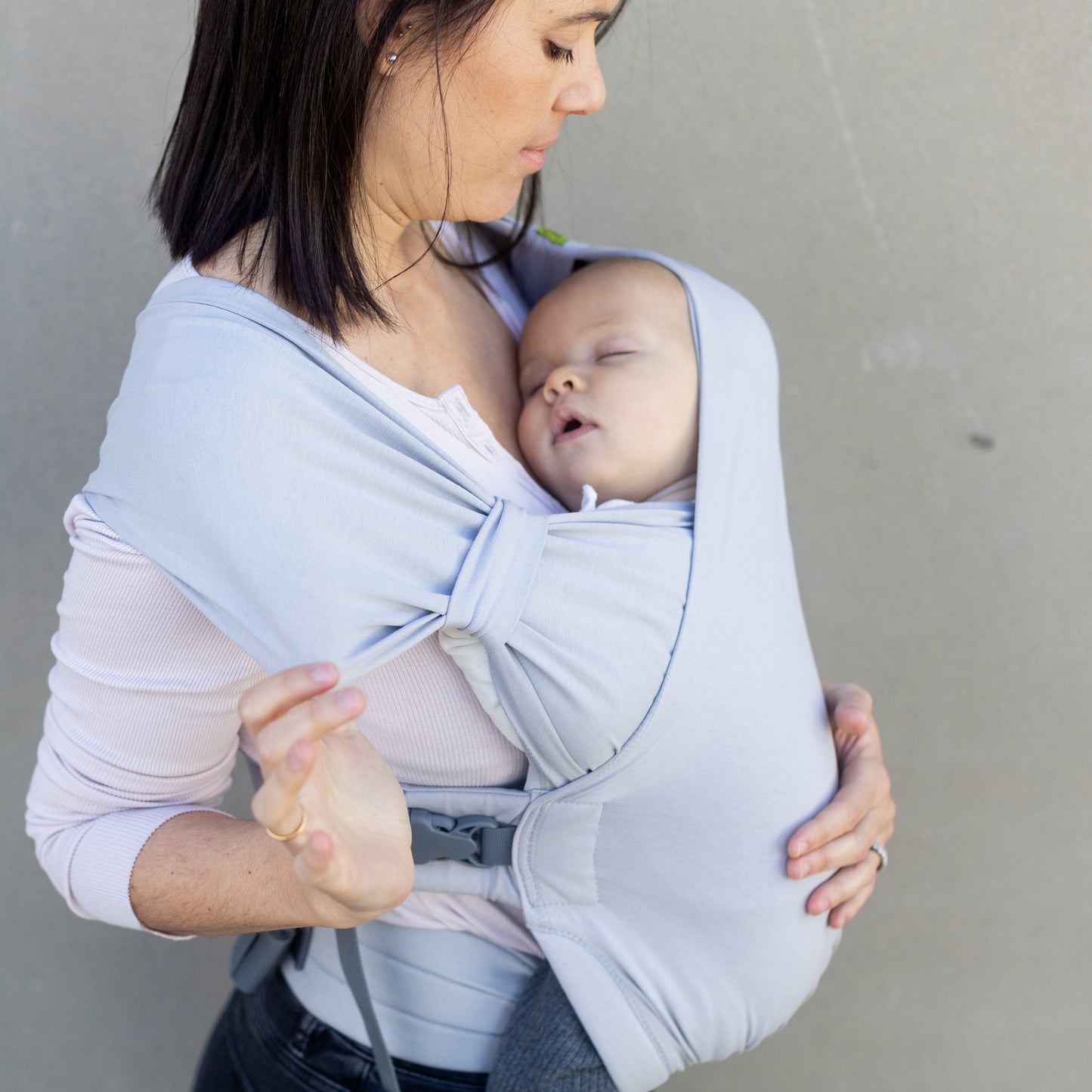 Woman holding a baby in a light gray baby carrier against a plain background