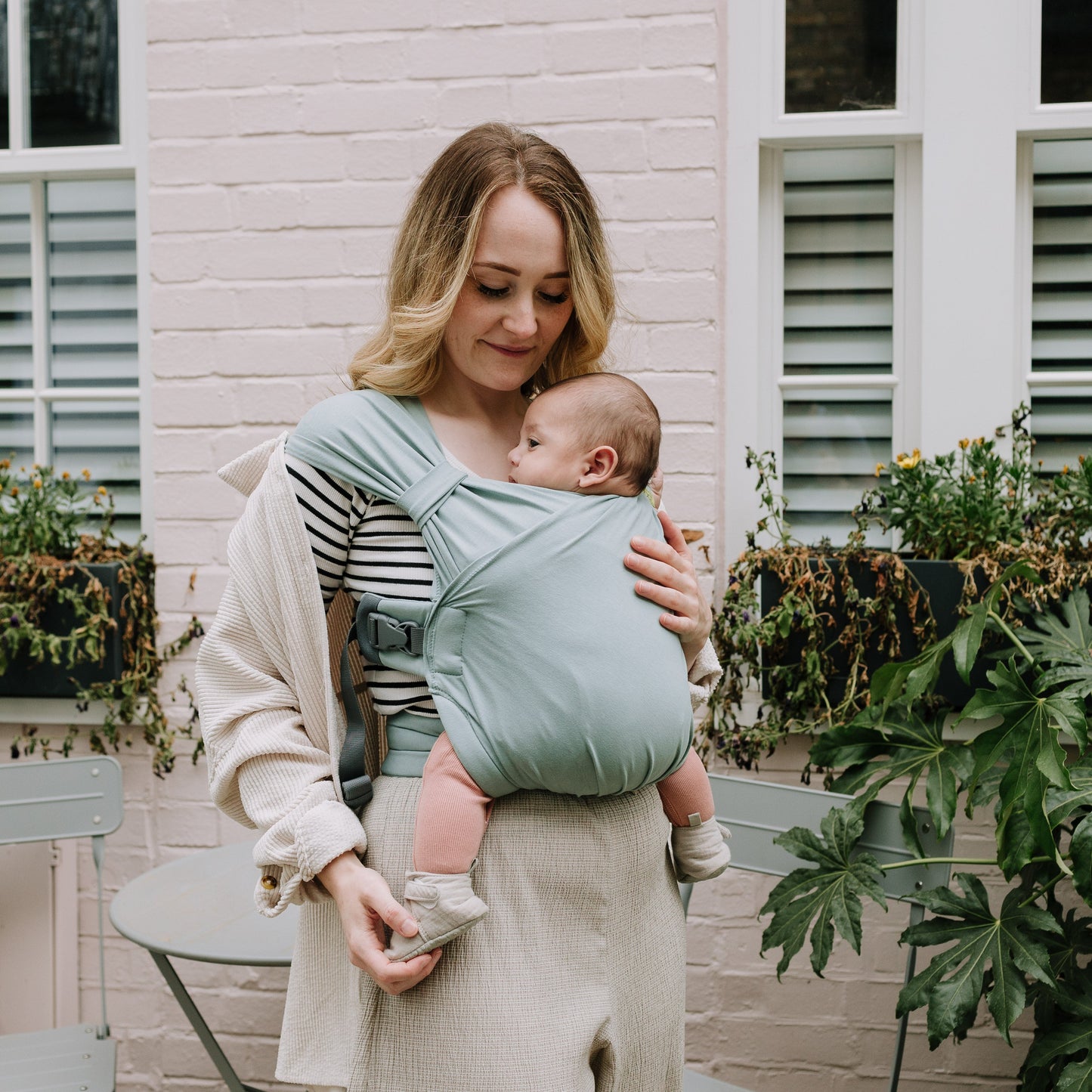 Woman holding a baby in a sling outdoors with plants and a building in the background
