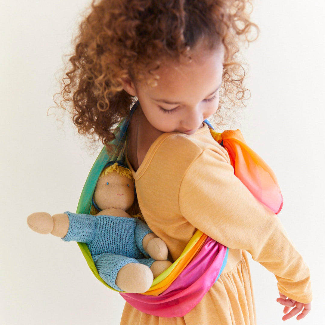 Child wearing a light peach dress carrying a doll with a rainbow silk scrarf tied to her body.