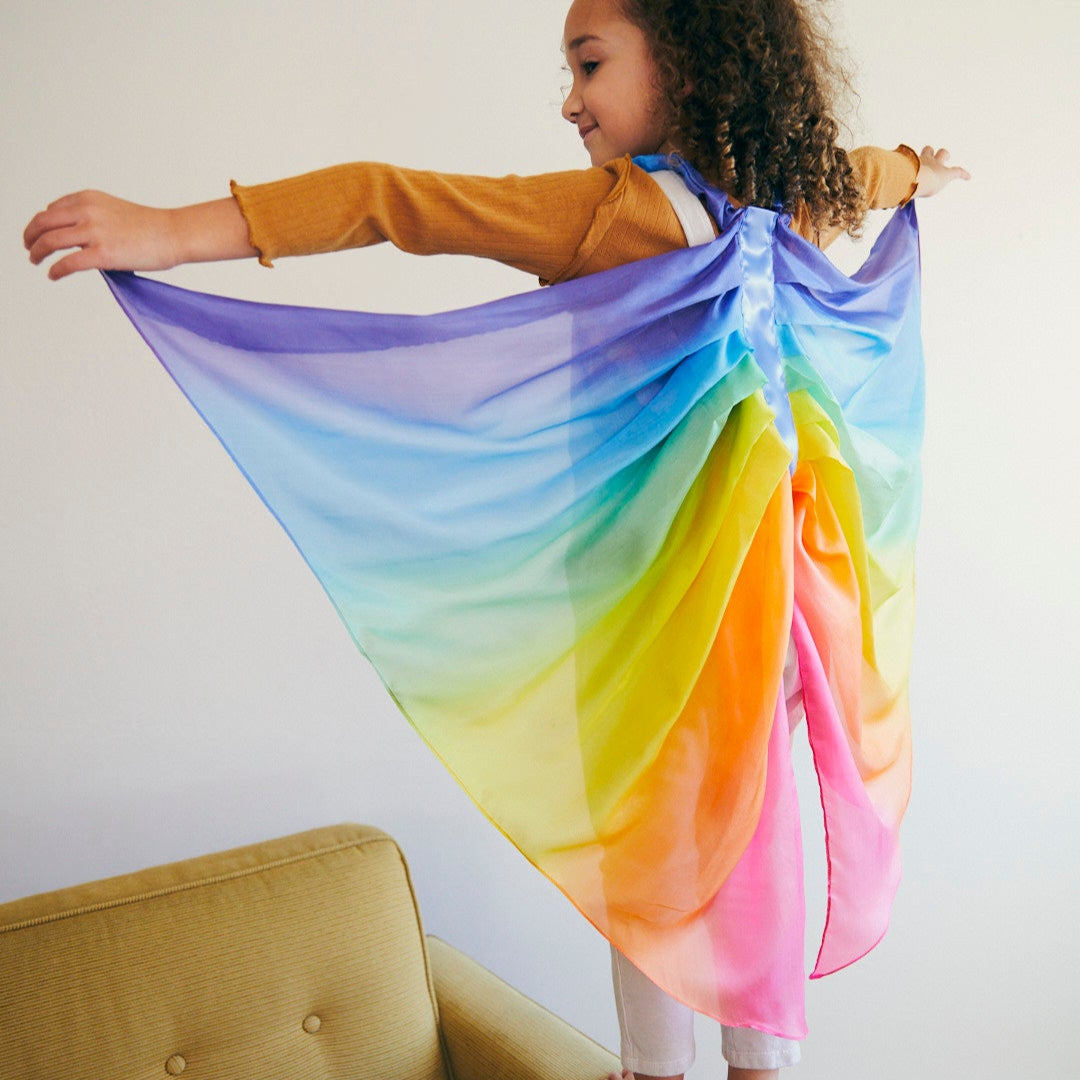 Child wearing a rainbow-coloured wings in a room with a beige couch.