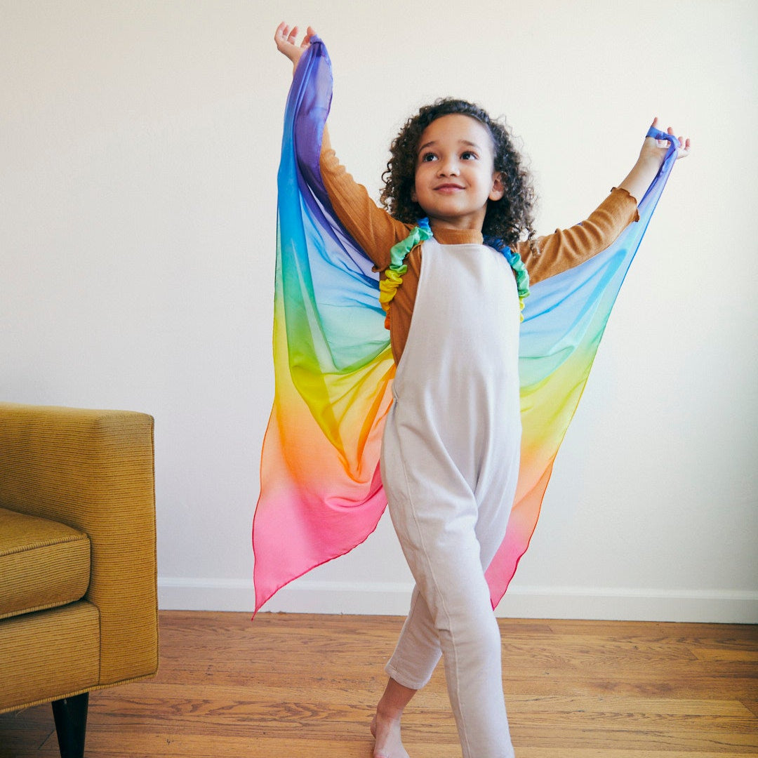 Child wearing a colourful rainbow wings indoors on a wooden floor.