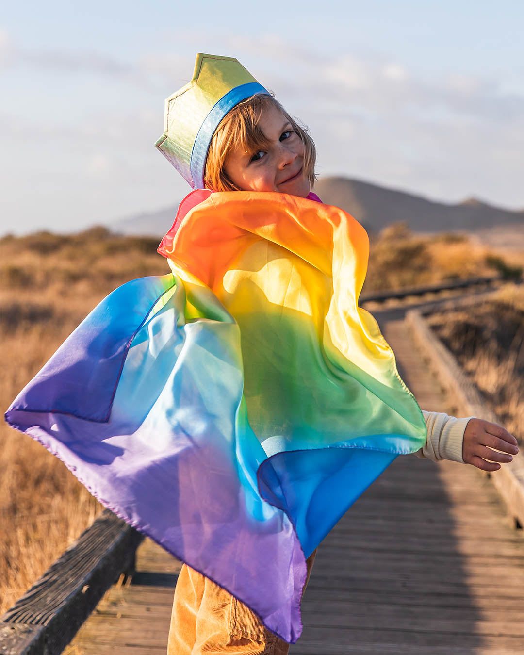 Child wearing a colourful rainbow cape on a wooden path with mountains in the background.
