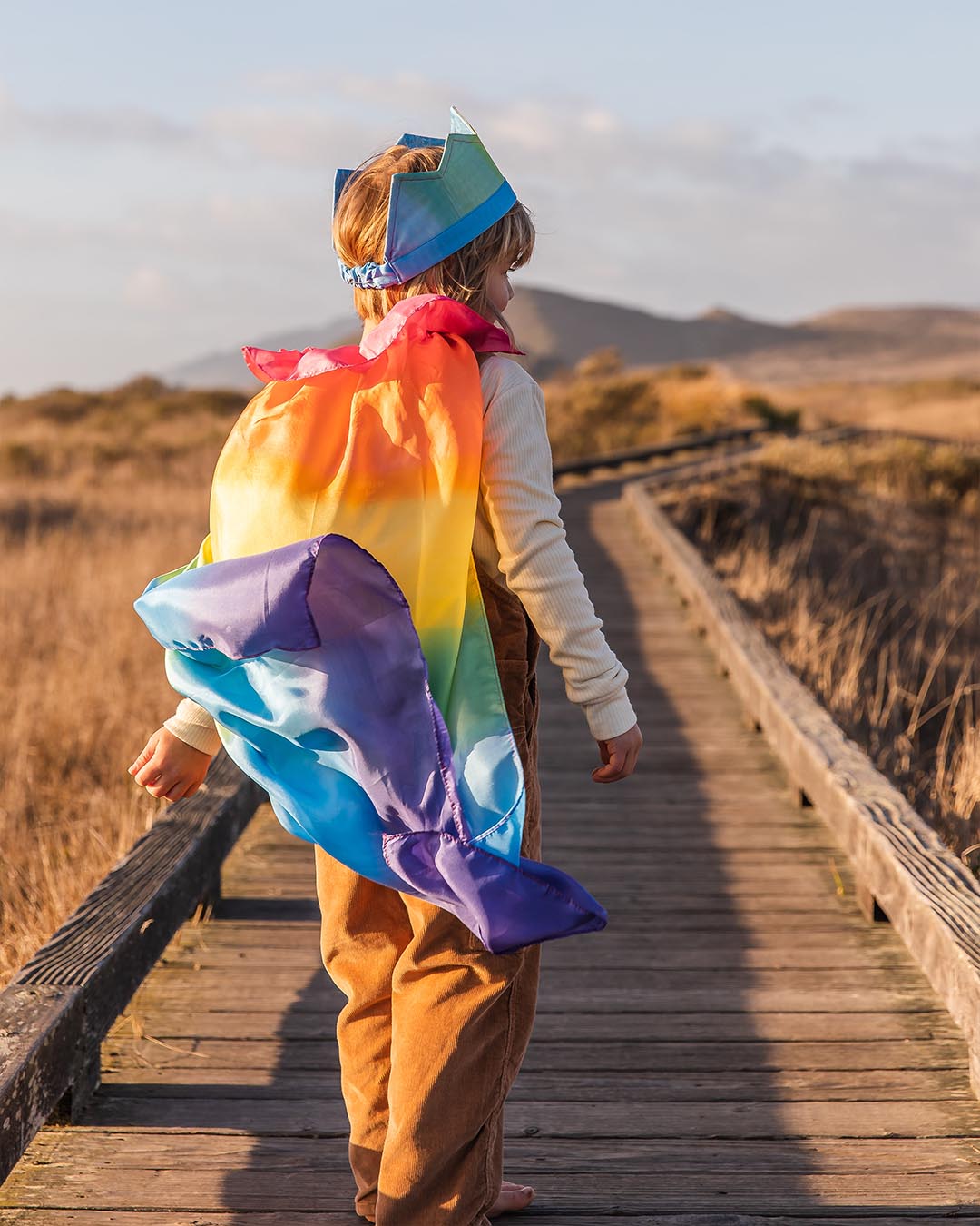Child wearing a rainbow cape on a wooden boardwalk with mountains in the background