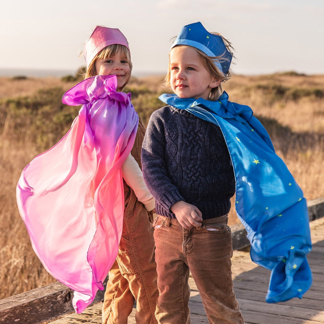 Two young children outdoors on a wooden boardwalk wearing colorful fabric capes and hats. The child on the left wears a pink and purple cape, and the child on the right wears a blue cape with small yellow star details and a blue hat.