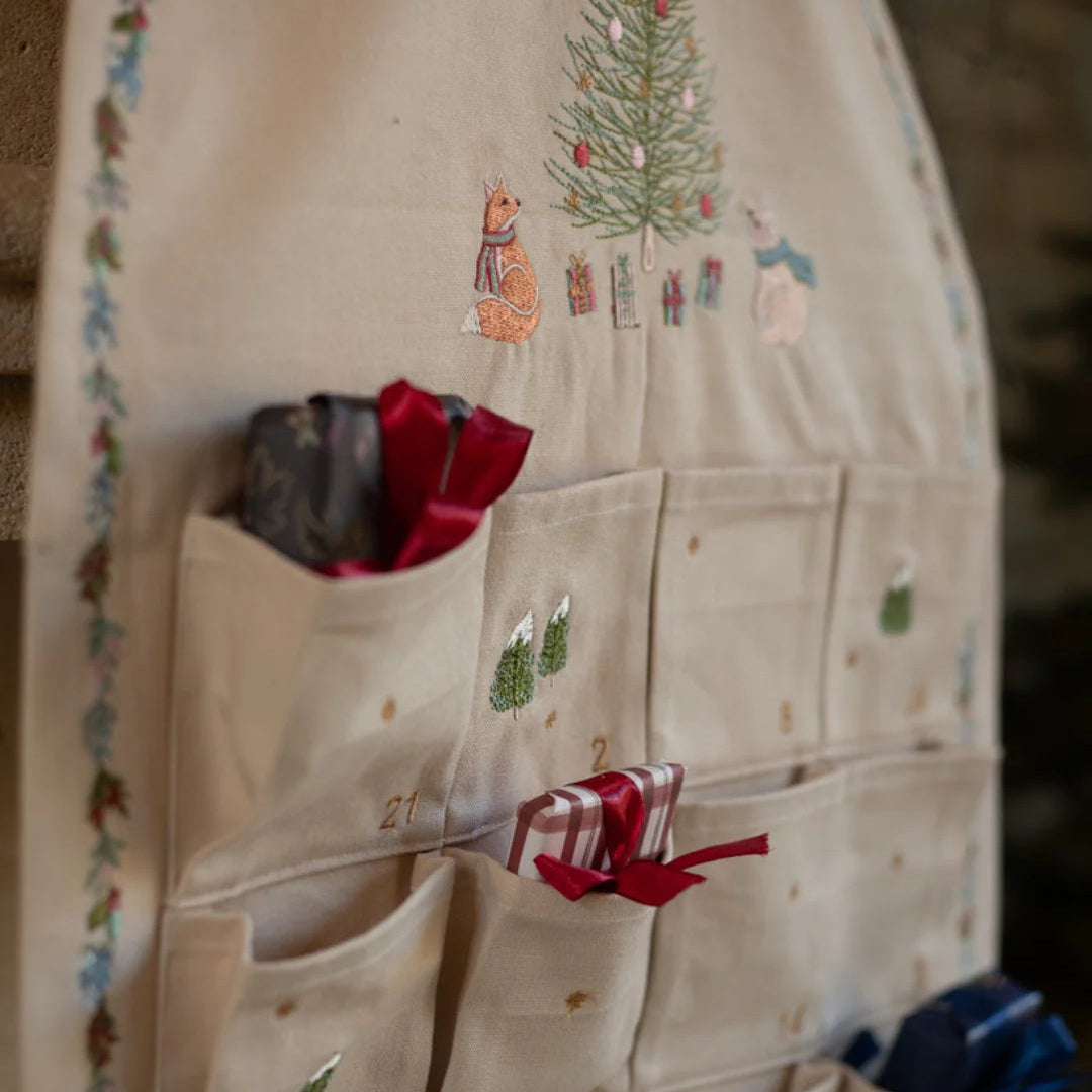 Cream advent calendar with embroidered Christmas scenes and red ribbons.