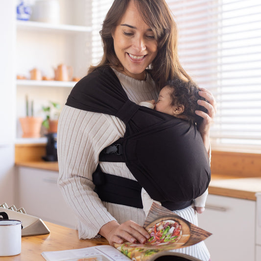 Woman reading a magazine while holding a baby in a sling at home.