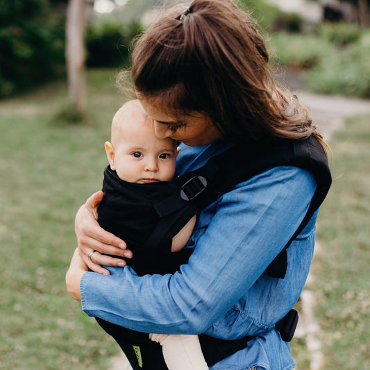 Woman carrying a baby in a black carrier outdoors on a grassy path.