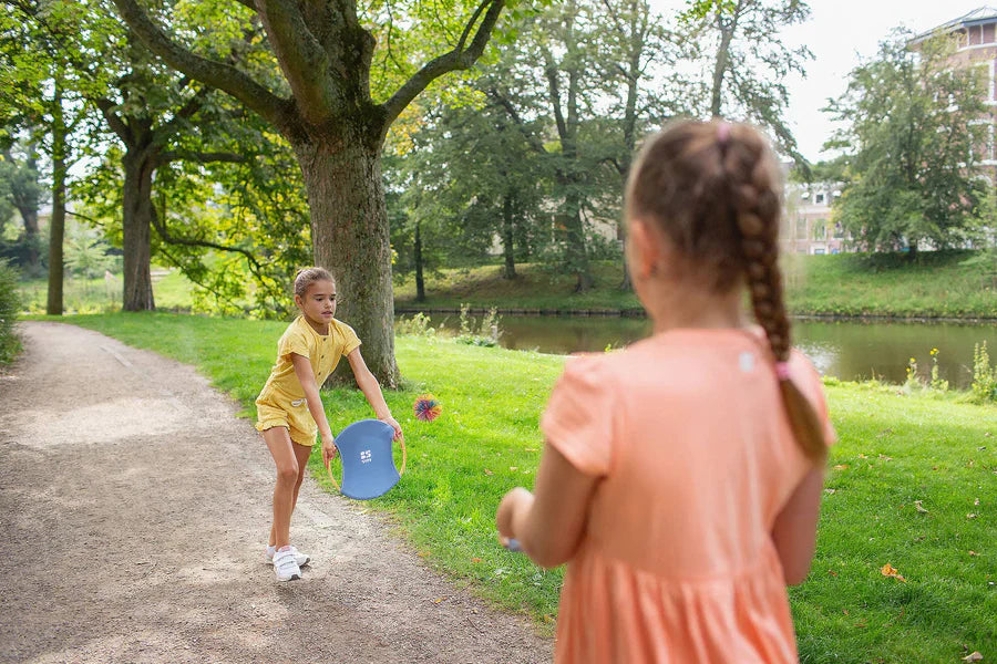 Two young girls playing with a blue bounce racket in a park.