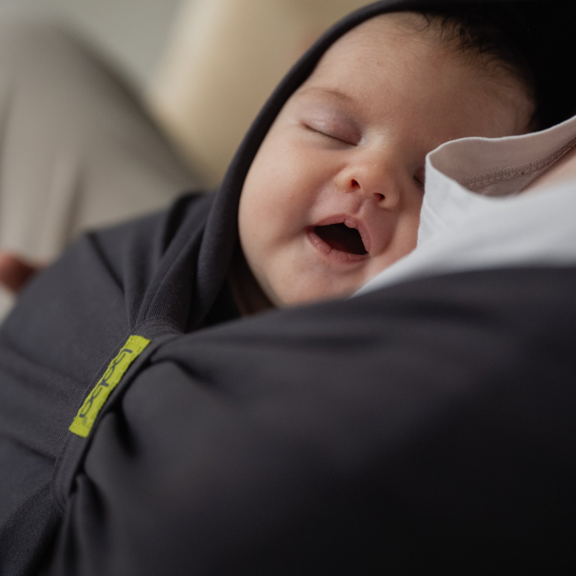 Baby sleeping in a dark gray swaddle with a yellow tag, held by an adult.