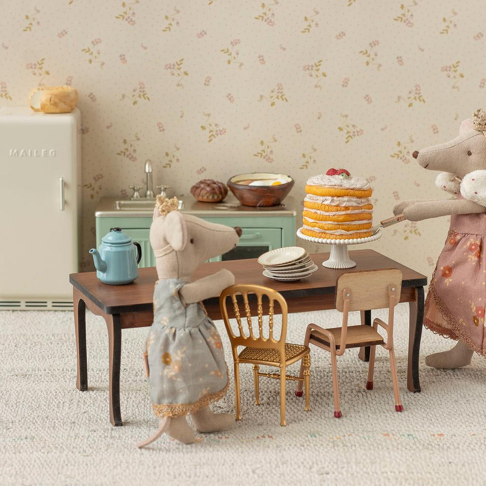 Two toy mice in dresses standing at a table with a cake and tea set, in front of a floral wallpapered wall.