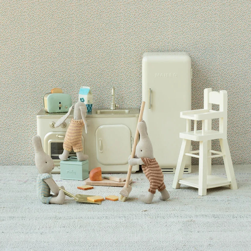 A miniature dollhouse kitchen scene with rabbit figurines, including one washing dishes at the sink, another standing on a stool near a toaster, and another holding a broom. The set includes a retro-style refrigerator, stove, high chair, and small accessories, arranged against a textured light gray wall and floor.