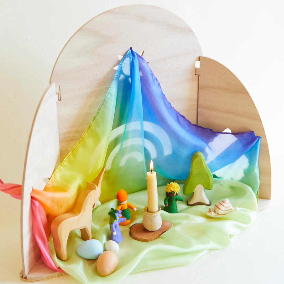 Seasonal table display with rainbow silk, toys, and a candle on a white background