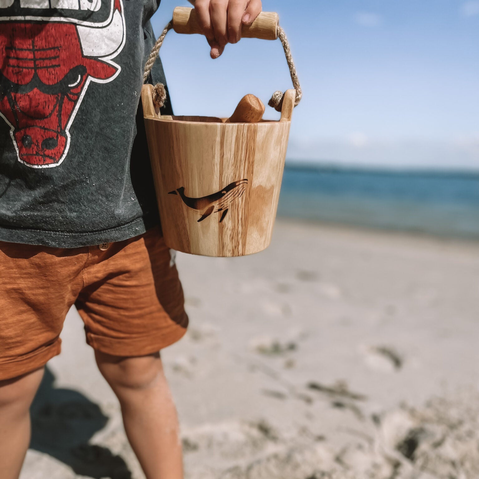 Child holding a wooden bucket on a sandy beach with ocean in the background