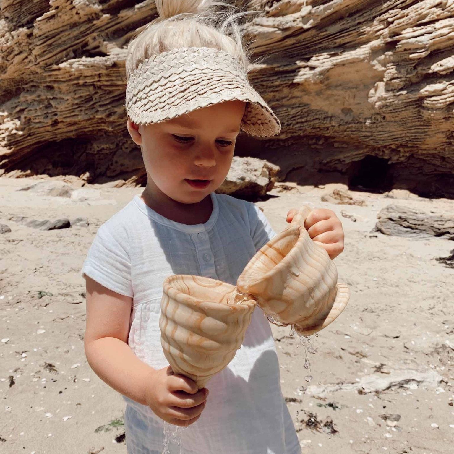 Child on a beach holding wooden toys wearing a visor and white dress.