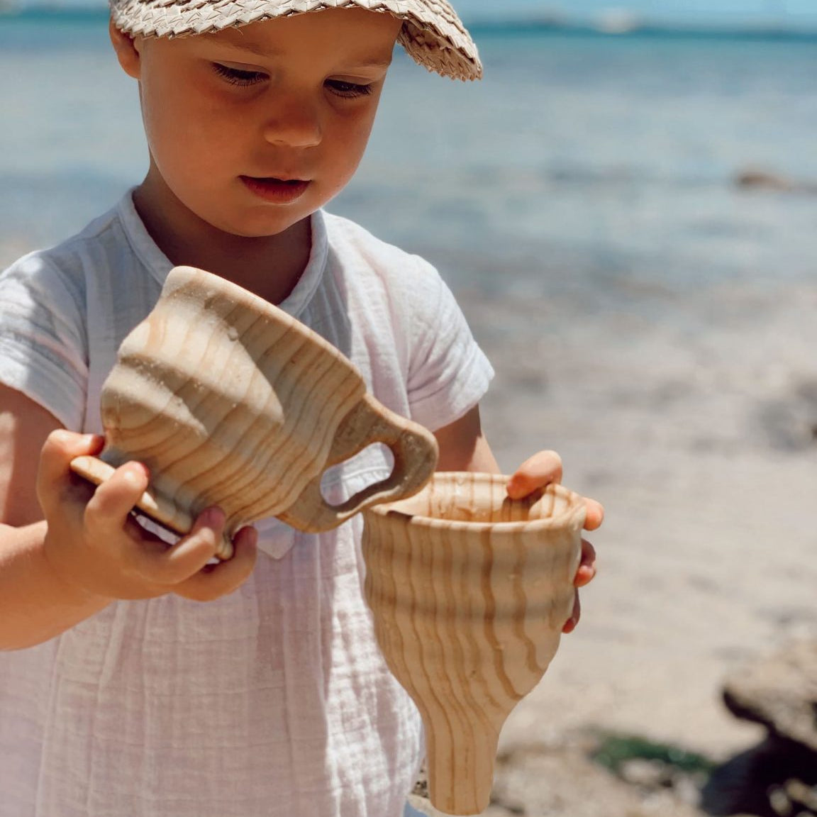 Child holding two wooden cups on a beach with ocean in the background