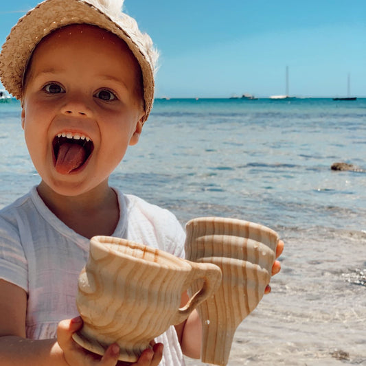 Child holding wooden funnel and cup on a beach with ocean and sky in the background