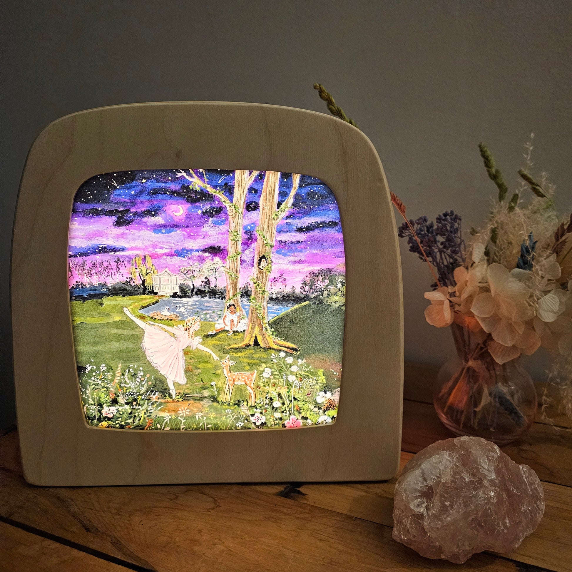 A wooden light box with rounded corners sits on a wooden table, displaying a colourful glowing scene. The box is styled on a rustic wooden surface next to a pink crystal and a vase of dried flowers against a dark gray wall.